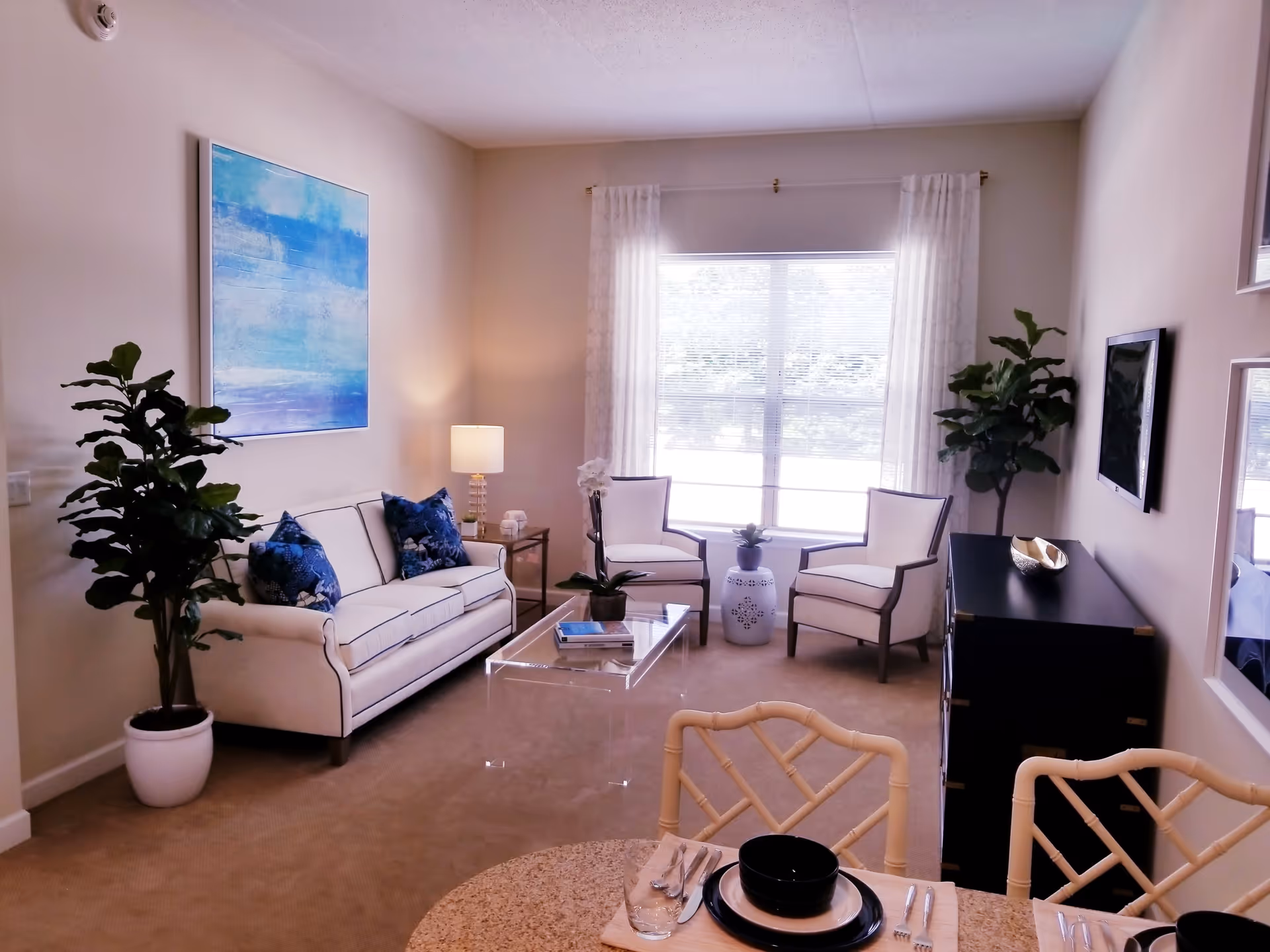 A bright living room with a white sofa adorned with blue pillows, two white armchairs, a clear glass coffee table, and a black cabinet with a decorative bowl. There are two large green plants in white pots, a blue abstract painting on the wall, and a large window with white curtains letting in natural light. In the foreground, part of a dining table set with black dishes and silverware is visible.