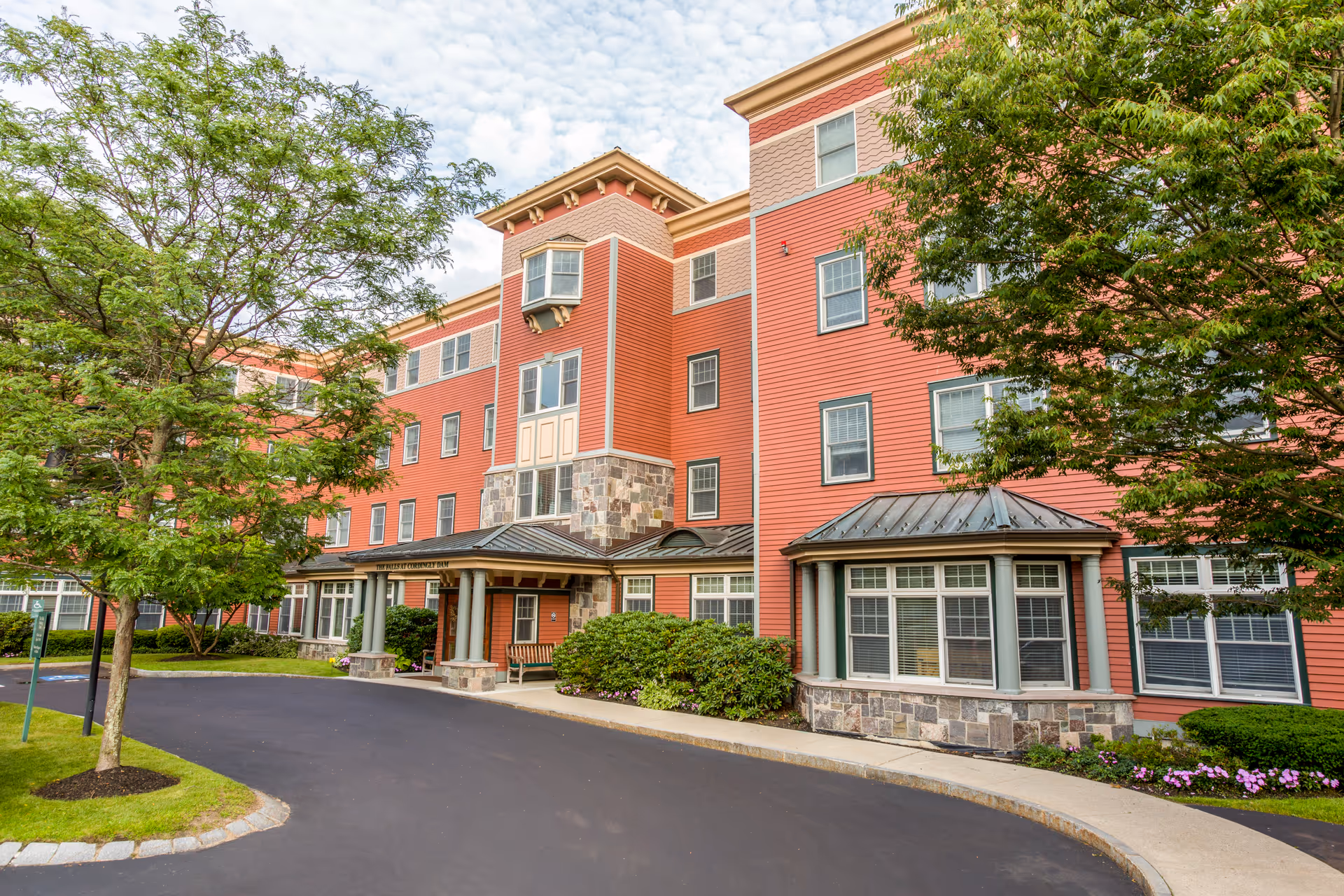 Front exterior of a red multi-story senior living building with a covered entrance, stone accents, and surrounding trees.