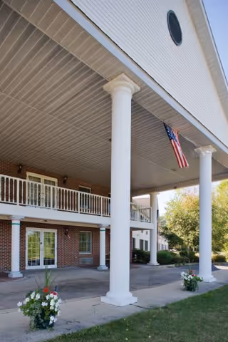 Exterior view of a senior living facility named Riverview Ridge showing a covered entrance with large white columns, a balcony with white railings, brick walls, an American flag hanging from the ceiling, and flower pots with white and red flowers on the ground near the entrance.