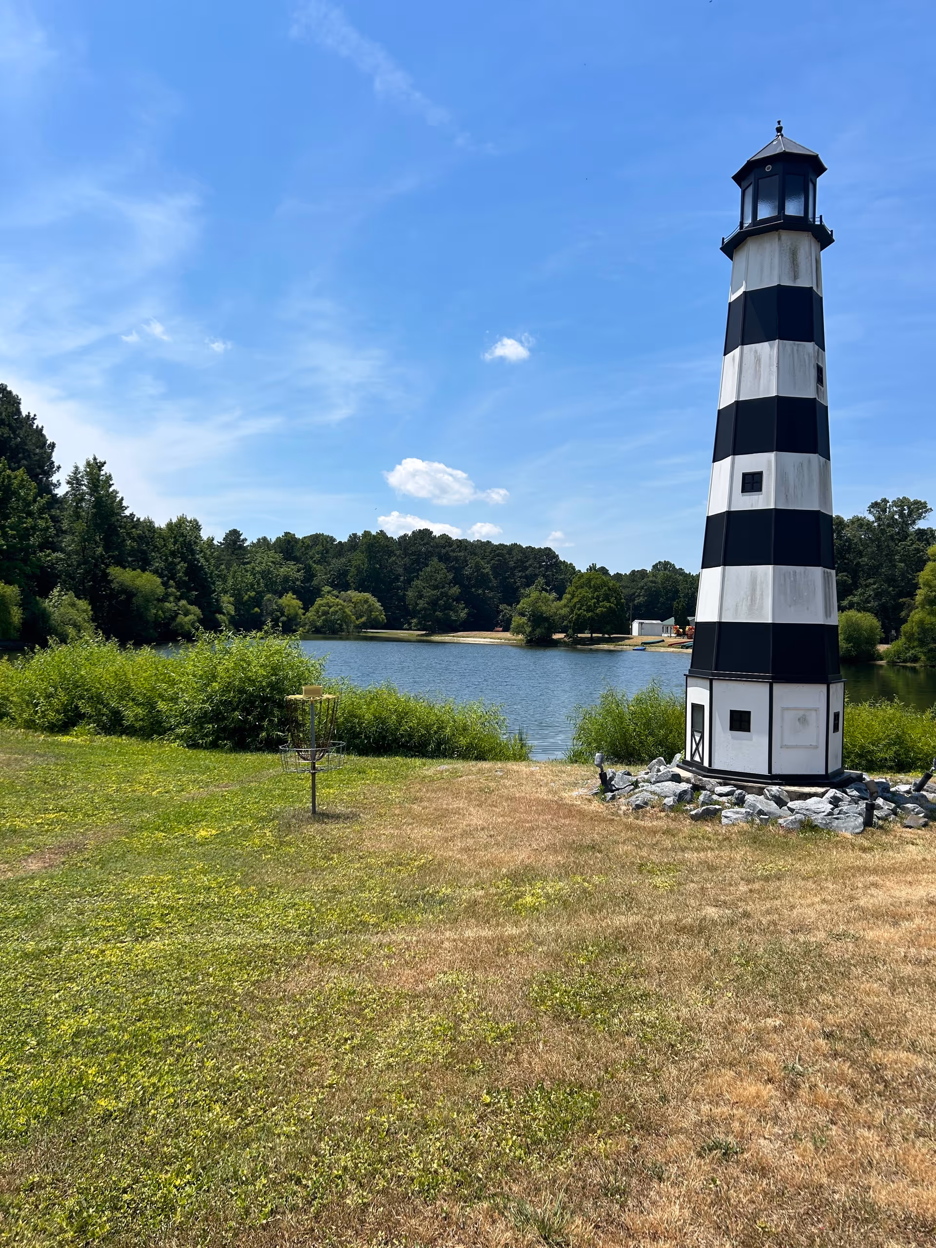 A black and white striped lighthouse structure stands on a grassy area near a body of water with trees in the background under a blue sky with some clouds.