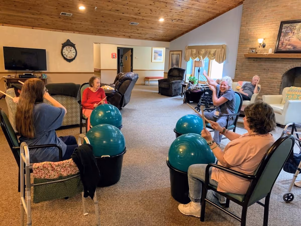 A group of elderly individuals seated in a living room area participating in a drumming exercise using large blue exercise balls and drumsticks. The room has a high wooden ceiling, carpeted floor, and a brick fireplace. Some people are actively drumming while others watch or relax in chairs.