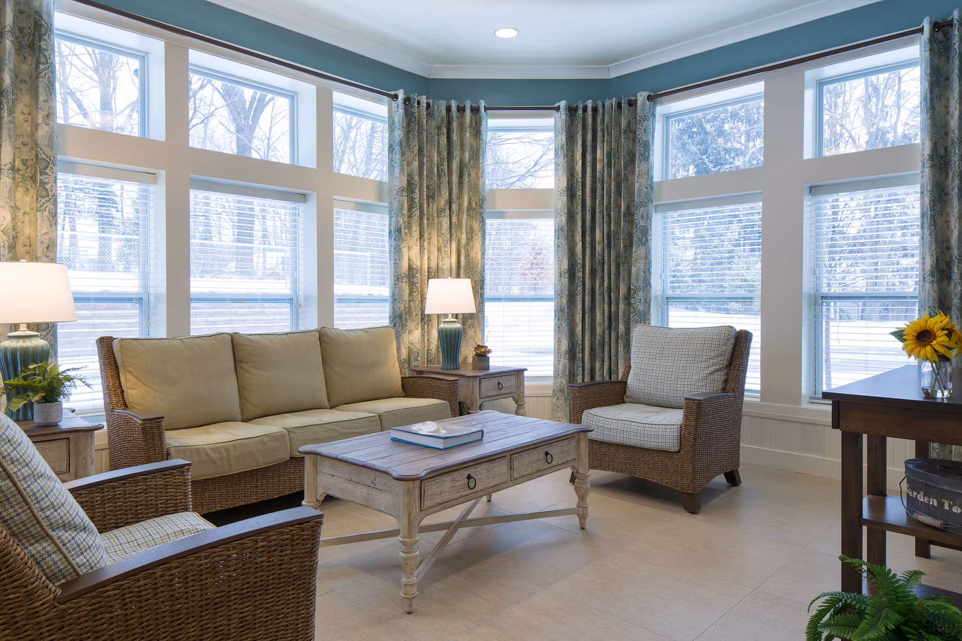 A bright and cozy living room with large windows letting in natural light. The room features a beige cushioned sofa, two armchairs with plaid cushions, a wooden coffee table with a book on it, two table lamps on side tables, floral curtains, and a wooden console table with a vase of sunflowers and a decorative basket underneath.