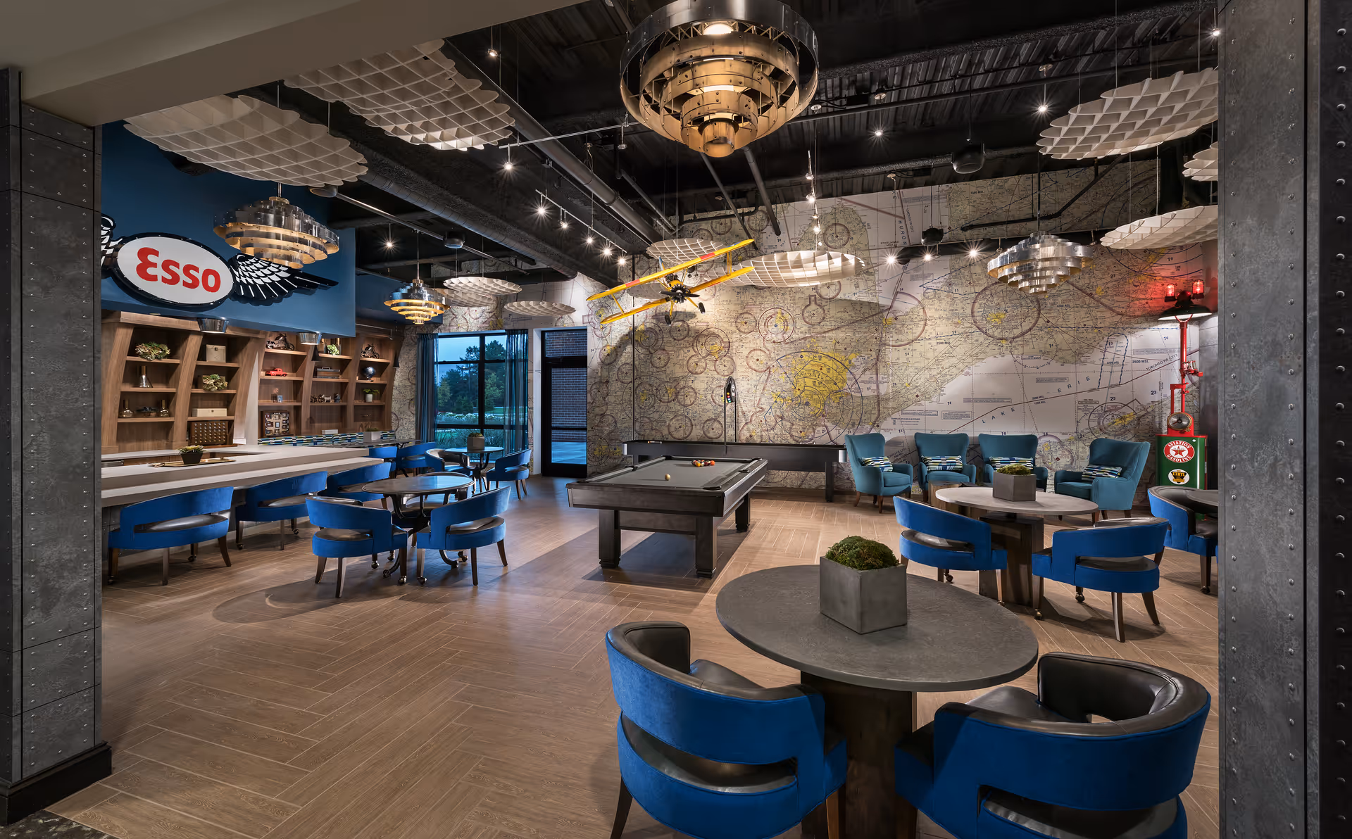 Spacious lounge with blue chairs and round tables, a pool table in the center, an 'Esso' wall sign, decorative ceiling lights, and a large map mural on the back wall.