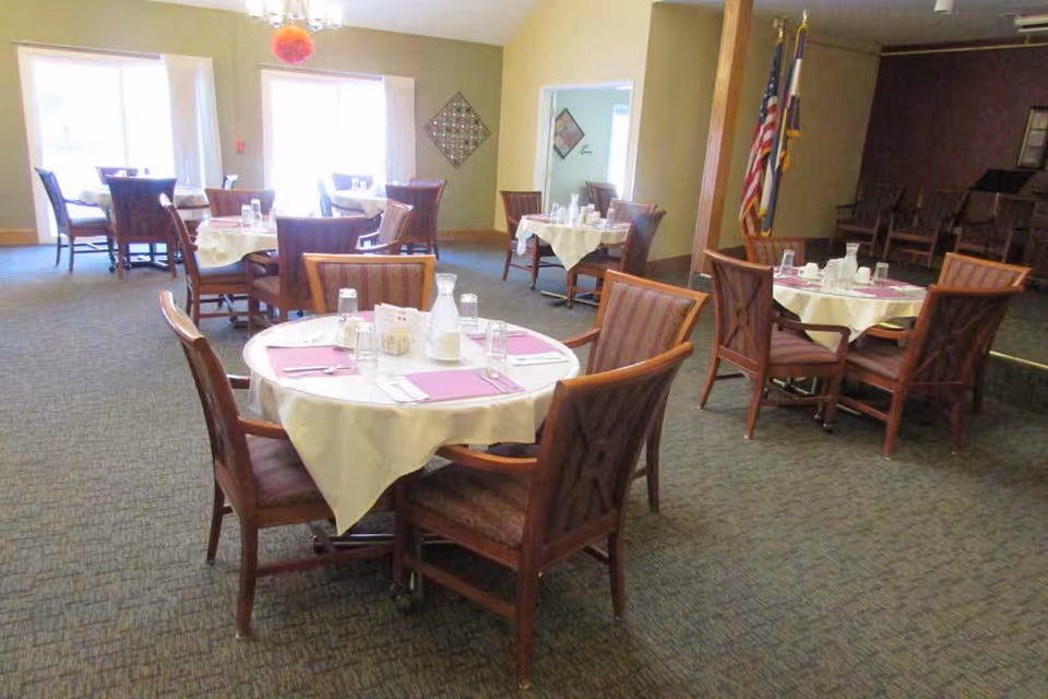 Dining room with several round tables set with placemats, glassware, and wooden chairs.