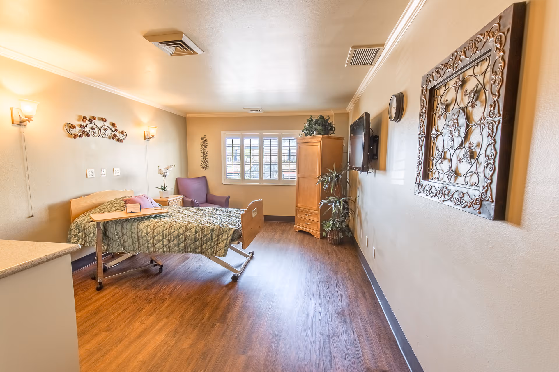 A bright and spacious bedroom in a senior living facility with a hospital-style bed covered in a green quilt, a purple armchair near a window with white shutters, a wooden cabinet, a wall-mounted TV, decorative wall art, and wooden flooring.
