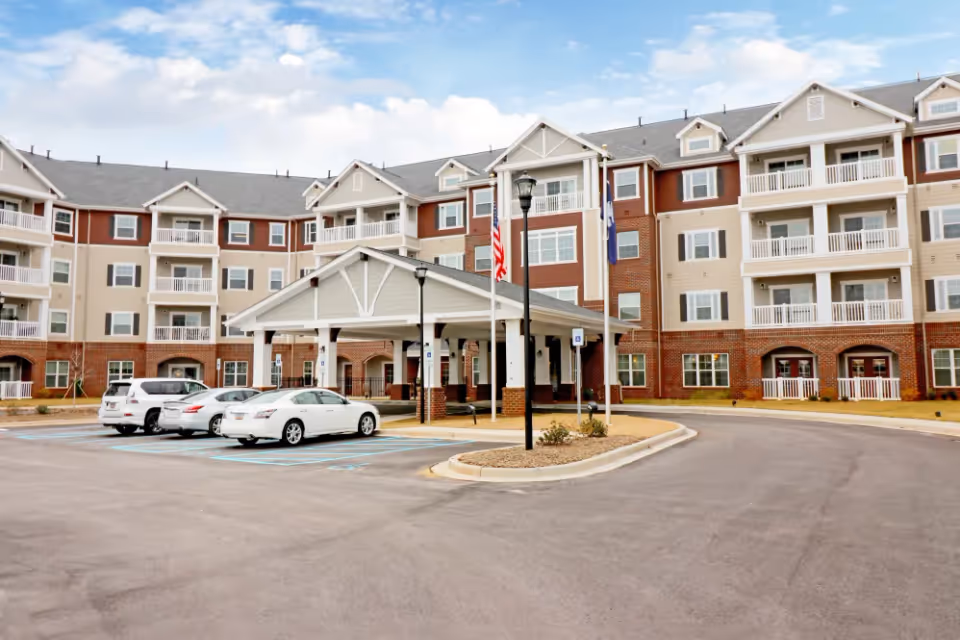 Exterior view of a large senior living facility building with multiple floors, balconies, and a covered entrance. Several cars are parked in front, and there are American and other flags near the entrance. The sky is partly cloudy.