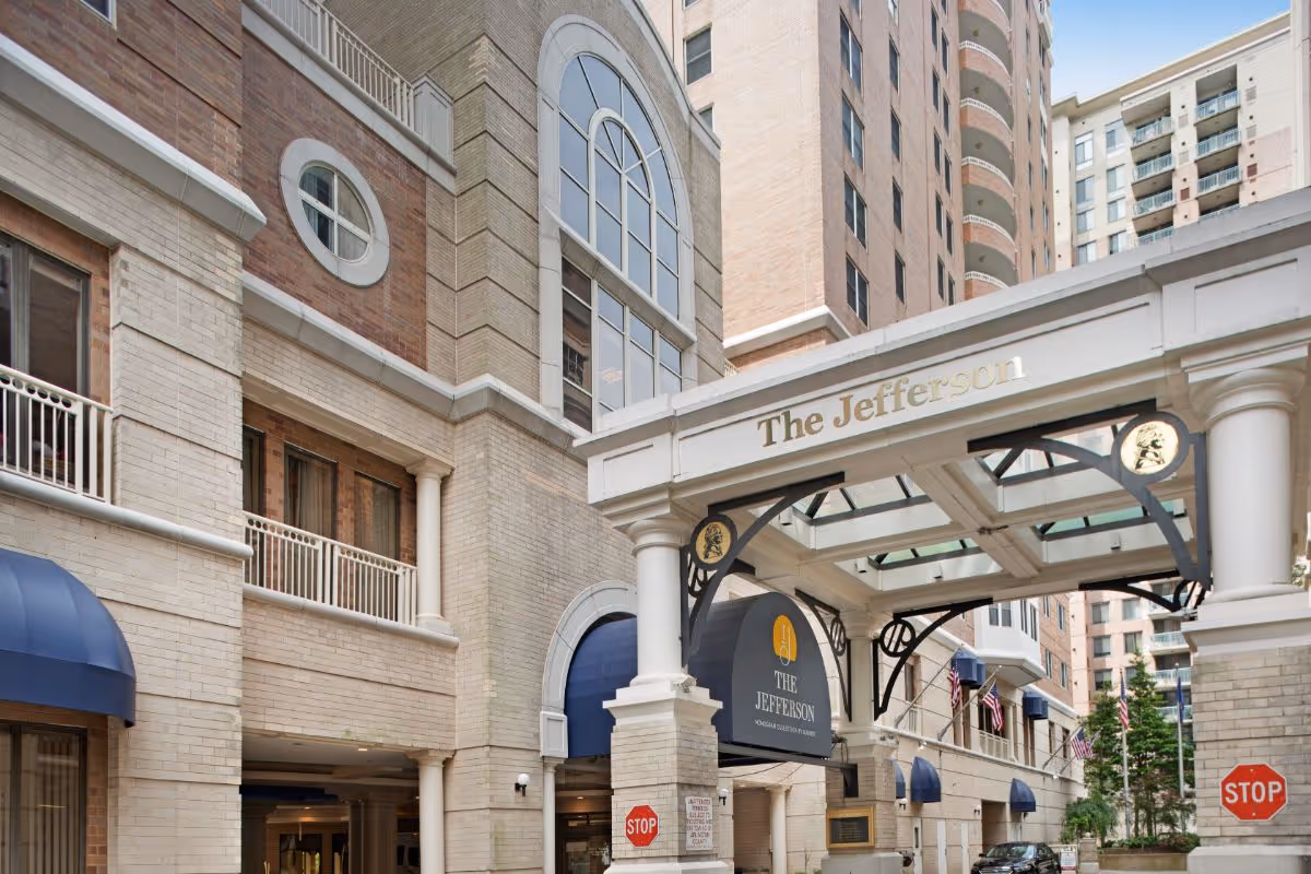 Exterior view of The Jefferson senior living facility entrance with a covered driveway supported by white columns, beige brick walls, blue awnings, and multiple American flags displayed along the building.