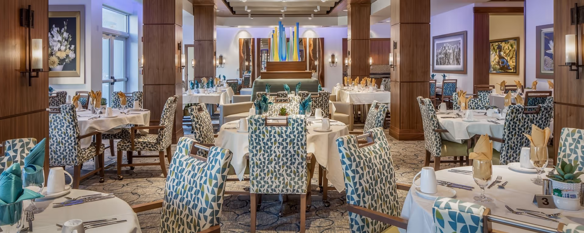 A spacious and elegantly decorated dining room with multiple round tables covered with white tablecloths, set with plates, glasses, cutlery, and folded napkins in teal and beige colors. The chairs have patterned upholstery in blue, green, and white. The room features wooden columns, wall sconces, framed artwork, and a decorative centerpiece with tall colorful glass vases in the background.