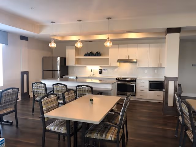 A modern kitchen and dining area in a senior living facility. The kitchen features white cabinets, a stainless steel refrigerator, oven, and microwave. Three pendant lights hang above a kitchen island with a white countertop. In front of the island, there are tables with plaid cushioned chairs arranged around them. The floor is dark wood, and the room is well-lit with natural light coming from a window on the left.