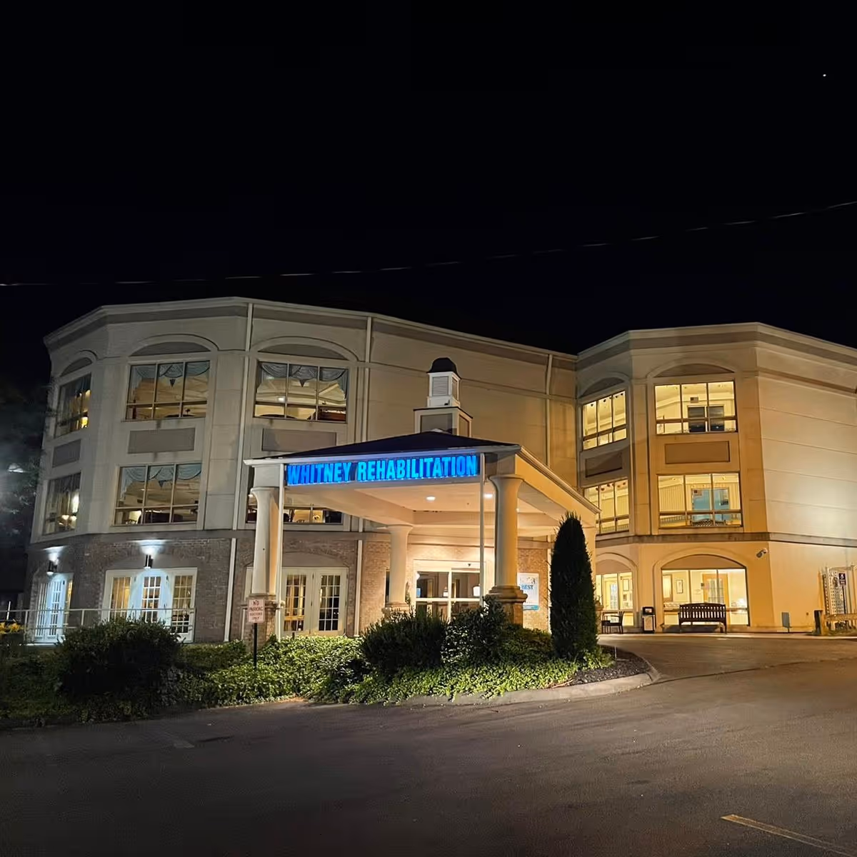 Night view of the Whitney Rehabilitation Care Center building with its entrance illuminated. The building has large windows and a covered drop-off area with a sign that reads 'WHITNEY REHABILITATION' in blue letters. There are bushes and a small tree near the entrance.
