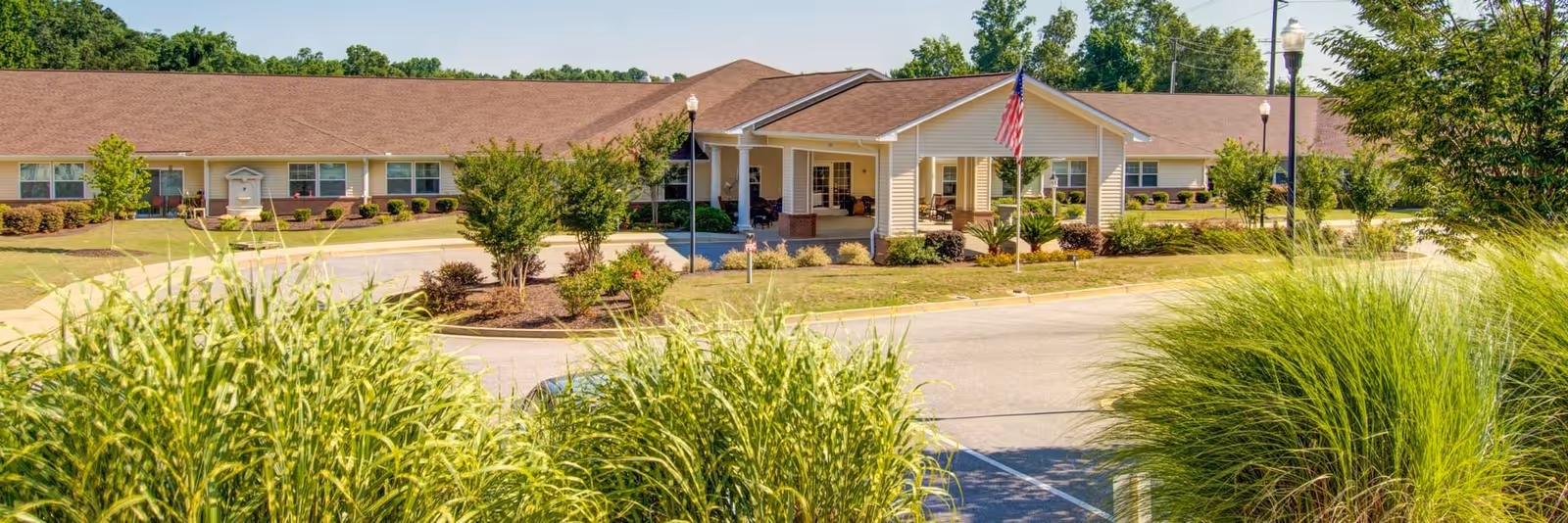 Exterior view of Spring Oak at Lexington, a single-story senior living facility with a brown roof and beige siding. The entrance features a covered porch with white columns and an American flag on a flagpole. The surrounding area includes well-maintained landscaping with bushes, trees, and ornamental grasses, along with a paved driveway and parking area.