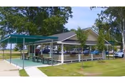 Outdoor covered picnic area with tables and benches next to a parking lot with several cars, surrounded by grass and trees under a clear sky.