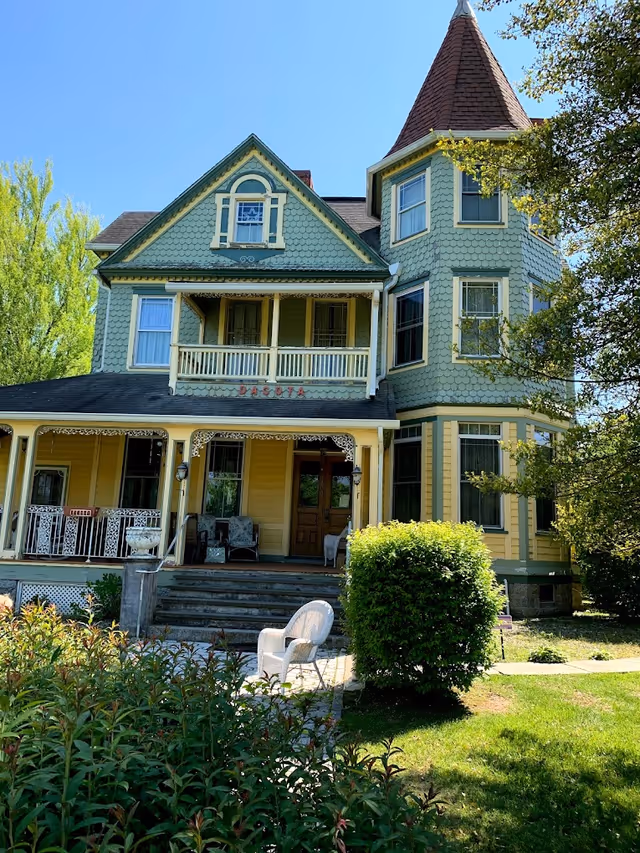 A large, charming Victorian-style house painted in green and yellow with a steeply pitched roof and a turret on the right side. The house has a covered porch with chairs and decorative trim. There is a white wicker chair on the lawn in front of the house, surrounded by bushes and trees under a clear blue sky.