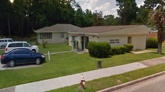 Exterior view of Tebeau House Personal Care Home, a single-story building with a small covered entrance, surrounded by green grass and bushes. There are three parked cars on the left side and a fire hydrant near the sidewalk in front.