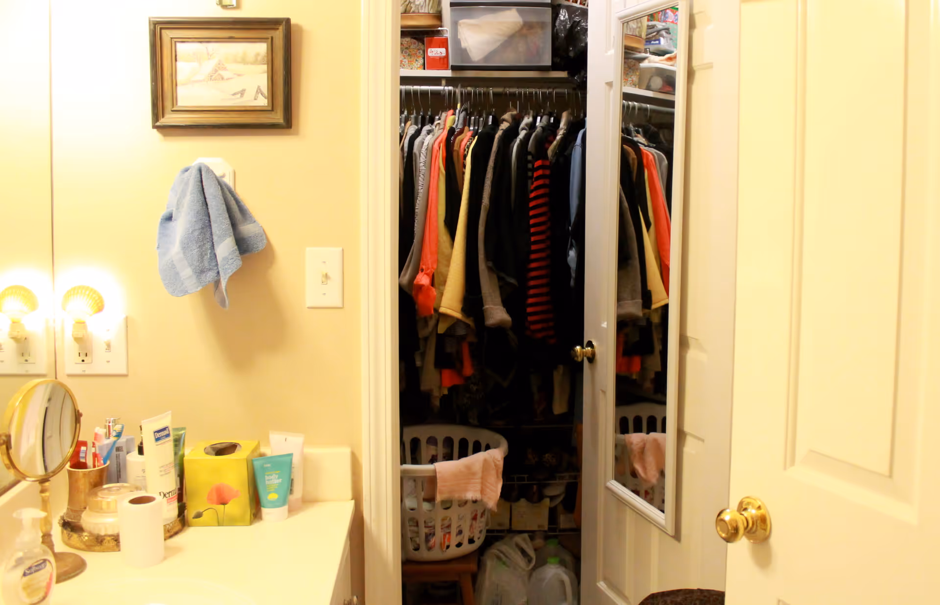 View of a bathroom vanity with various toiletries, a towel hanging on the wall, and a mirror reflecting a closet filled with hanging clothes and a laundry basket.