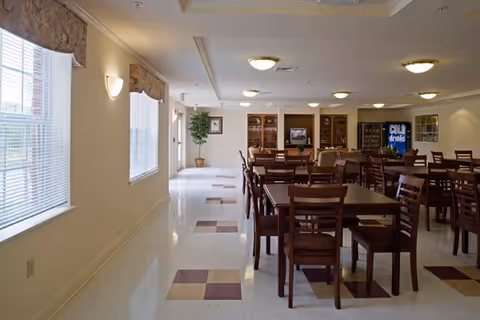 Spacious communal dining room with multiple wooden tables and chairs, windows along the left wall, and a vending machine area at the back.