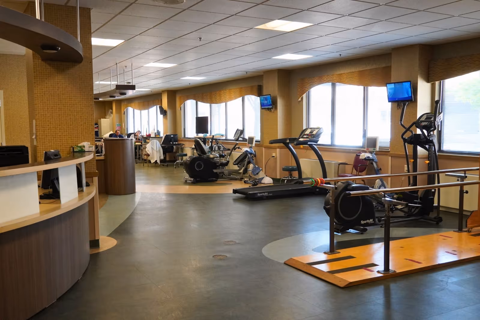 Interior view of a senior living facility fitness and rehabilitation area with exercise equipment including treadmills, stationary bikes, and parallel bars. There is a reception desk on the left and large windows on the right letting in natural light.