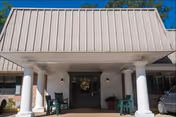 Entrance of Harvest Manor Healthcare and Rehabilitation featuring a covered porch with white columns, two green rocking chairs, potted plants, and a decorative wreath on the front door under a clear blue sky.