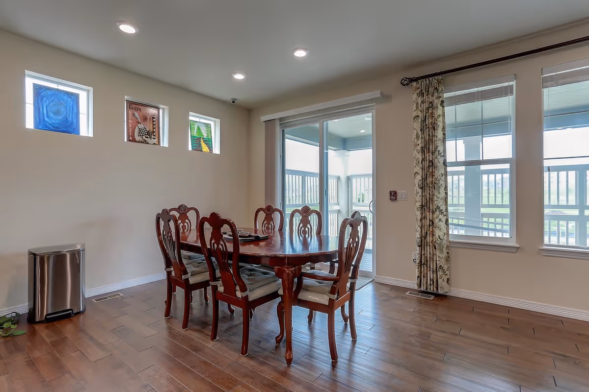 A dining room with a wooden table and six matching chairs on hardwood floors, sliding glass doors to a patio, and windows along the wall.