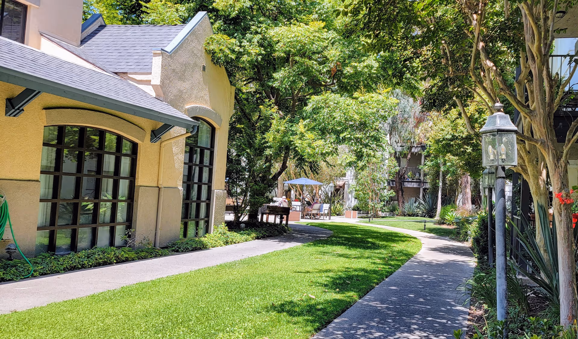 A sunny outdoor garden area at Westminster Terrace Senior Living featuring a curved concrete pathway surrounded by green grass and trees. On the left, there is a beige building with large arched windows, and on the right, there are lamp posts and more trees providing shade. In the background, there are outdoor seating areas with umbrellas.
