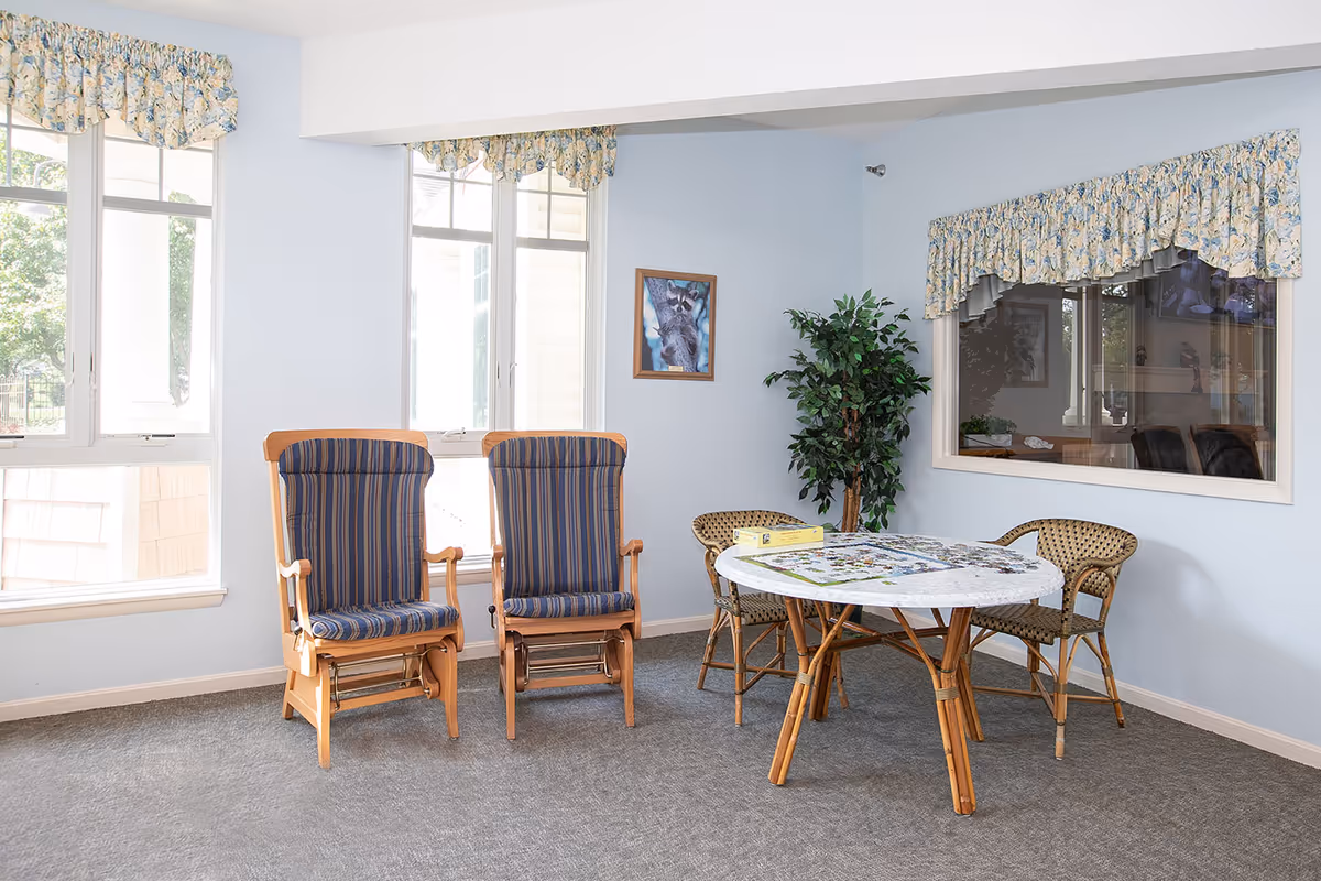 A bright common room with two upholstered wooden rocking chairs, a round table surrounded by wicker chairs with a puzzle on top, a potted plant and large windows.