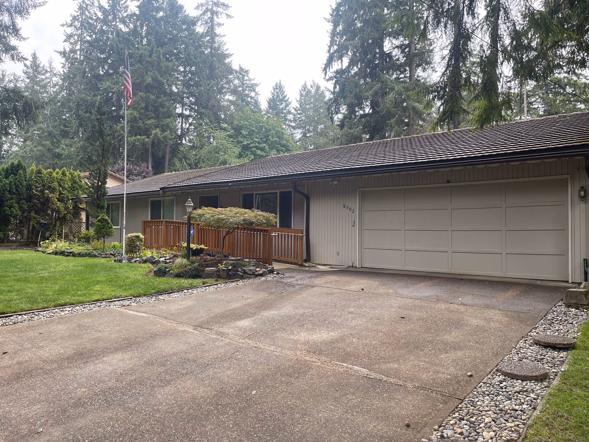 Exterior view of a single-story residential building with a large driveway, a two-car garage, a wooden ramp leading to the front door, a well-maintained lawn, and tall trees in the background. An American flag is mounted on a flagpole near the entrance.