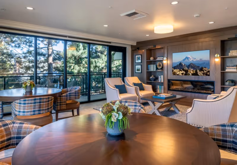 A cozy senior living common area with round wooden tables and plaid upholstered chairs in the foreground. In the background, there are beige armchairs with blue cushions arranged around a coffee table in front of a built-in wooden wall unit featuring a flat-screen TV displaying a mountain scene and a modern electric fireplace. Large glass sliding doors reveal a view of green trees outside.