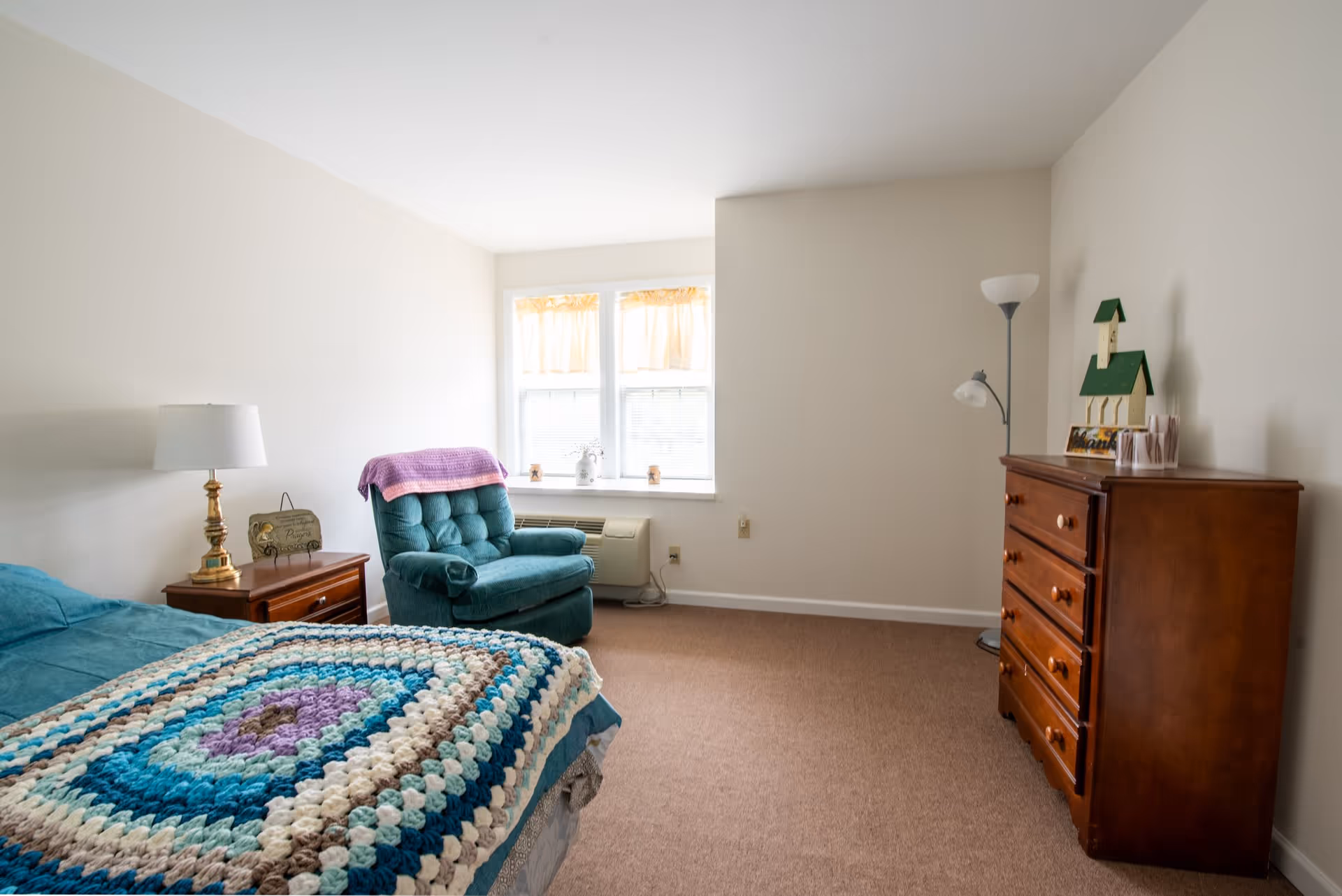 A simple bedroom with a bed covered by a multicolored crocheted blanket, a wooden nightstand with a lamp and a decorative sign, a teal upholstered armchair with a purple throw draped over it, a window with yellow curtains, a wooden dresser with decorative items on top, and a floor lamp in the corner.