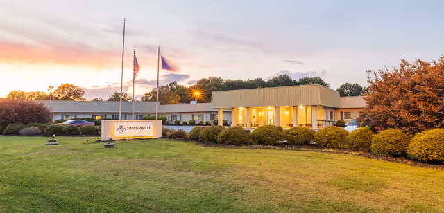 Exterior view of Simpsonville Post-Acute facility at dusk with a well-maintained lawn, bushes, two flagpoles with flags, and the building illuminated by warm lights.