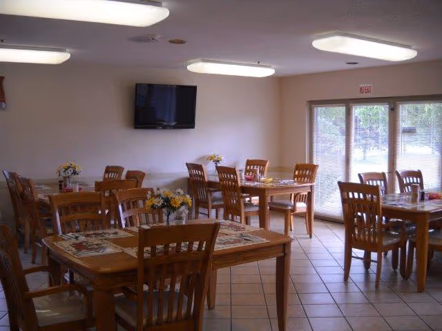A dining room with multiple wooden tables and chairs arranged neatly. Each table has a floral centerpiece and placemats. There is a wall-mounted flat-screen TV and large windows with blinds letting in natural light. The floor is tiled and the ceiling has rectangular fluorescent lights.
