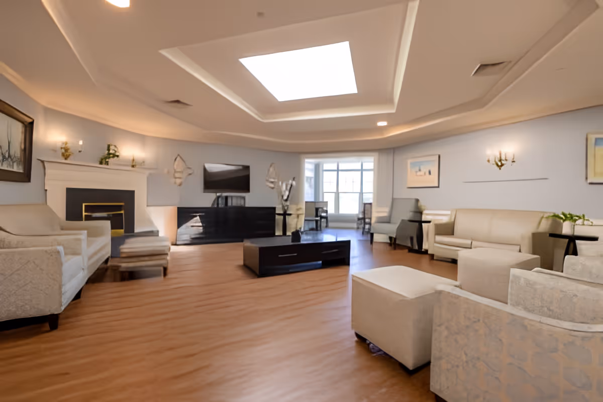 A spacious living room with beige sofas and armchairs arranged around a dark wooden coffee table. The room features a fireplace with decorative sconces on the wall above it, a flat-screen TV mounted on a black cabinet, and a large skylight in the ceiling. There are framed artworks on the light blue walls and a windowed area with additional seating in the background.