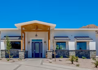 Front exterior view of a modern single-story building with a wooden entrance canopy, stone accents on the lower walls, large windows, and a clear blue sky above. Small desert landscaping with rocks and plants is visible in front of the building.