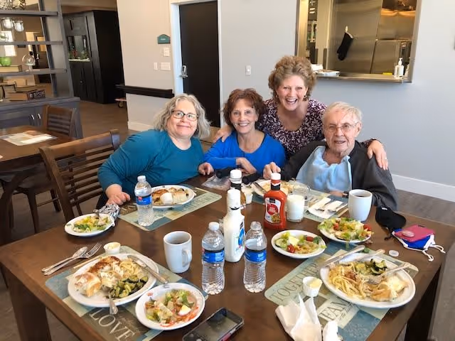 Four smiling seniors seated at a dining table in a communal dining room with plates of food, water bottles, and condiments.