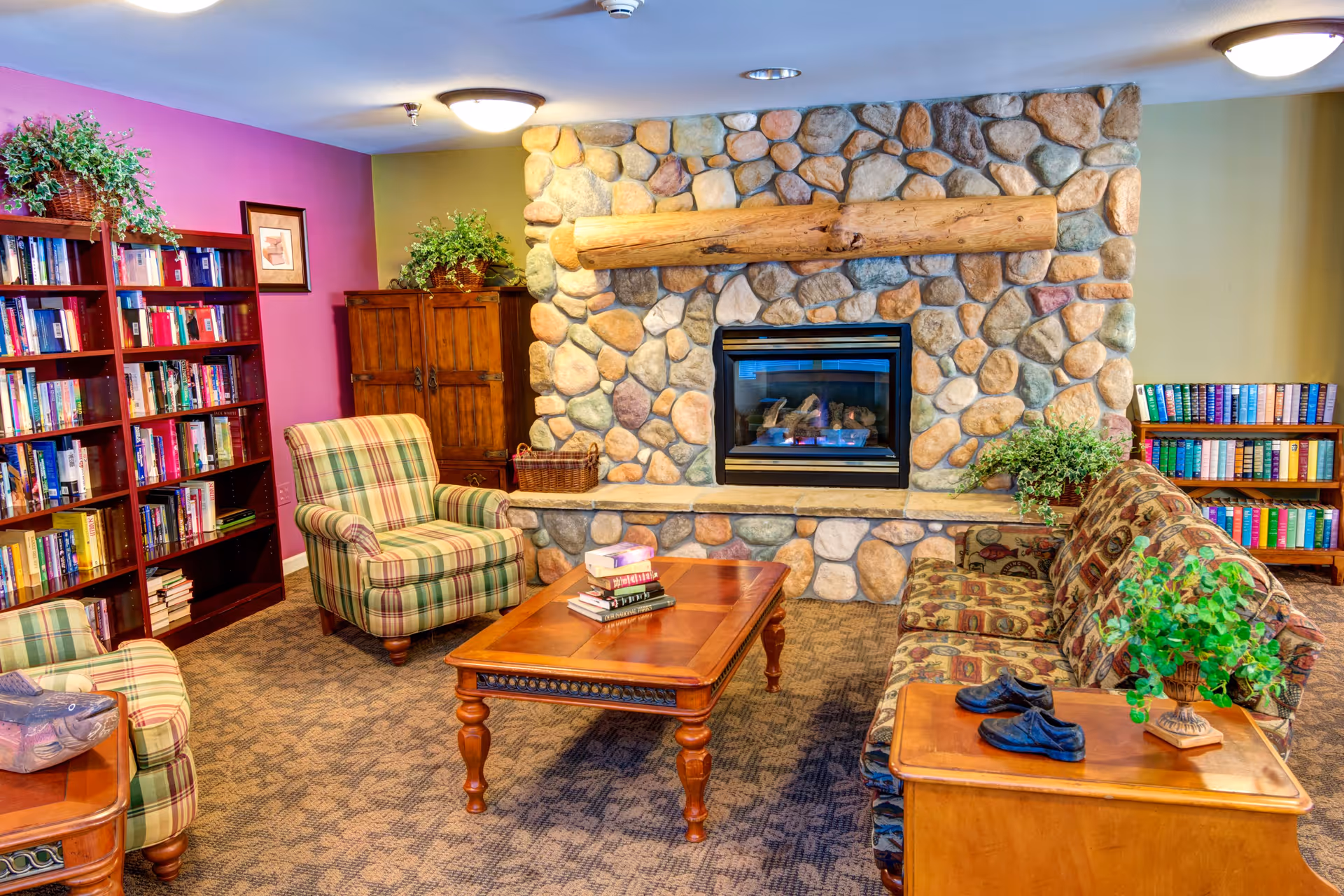 Cozy living room with a stone fireplace featuring a large wooden mantel. The room has a patterned carpet, a wooden coffee table with books, a plaid armchair, a patterned sofa, and wooden bookshelves filled with books. There are also green plants placed on furniture around the room.