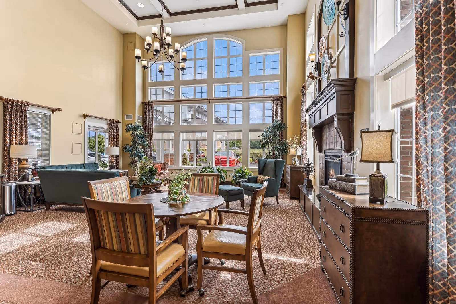 A bright and spacious living room area with large floor-to-ceiling windows allowing natural light to fill the space. The room features a round wooden table with four striped cushioned chairs, green upholstered armchairs, a fireplace with decorative plates above it, and various plants and lamps. The carpet has a patterned design, and the walls are painted a warm beige color.
