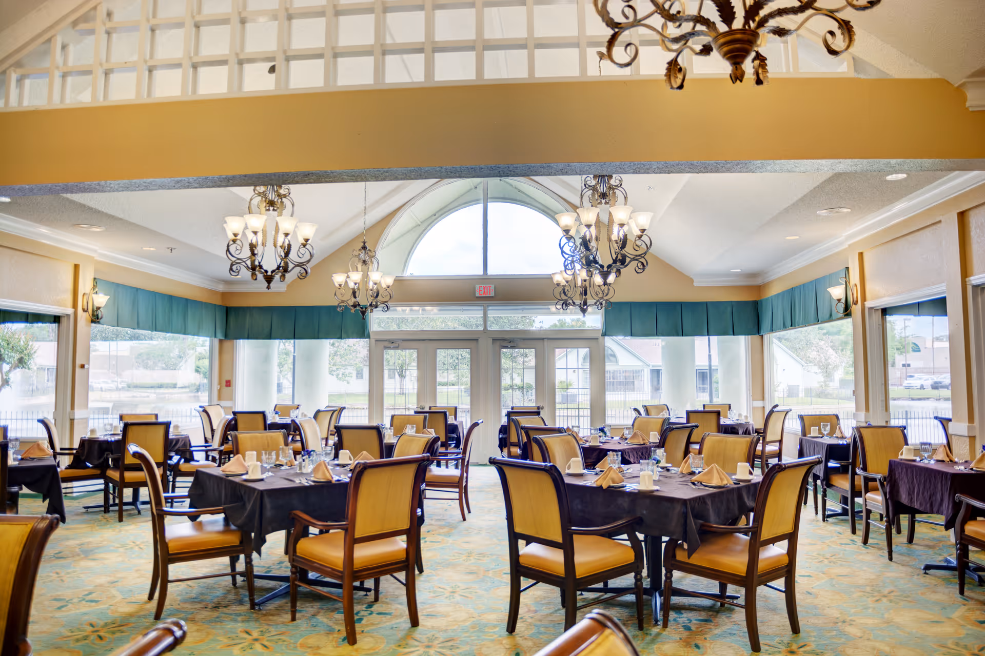 A bright and spacious dining room with multiple tables covered in dark tablecloths, each set with napkins, glasses, and silverware. The room features large windows with green valances, elegant chandeliers hanging from the ceiling, and a patterned carpet.
