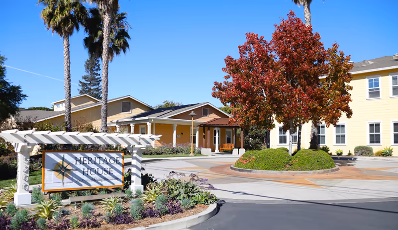 Exterior view of Heritage House, an assisted living community, showing a yellow building with multiple windows, a landscaped roundabout with a tree and bushes, palm trees, and a sign with the facility's name in front.