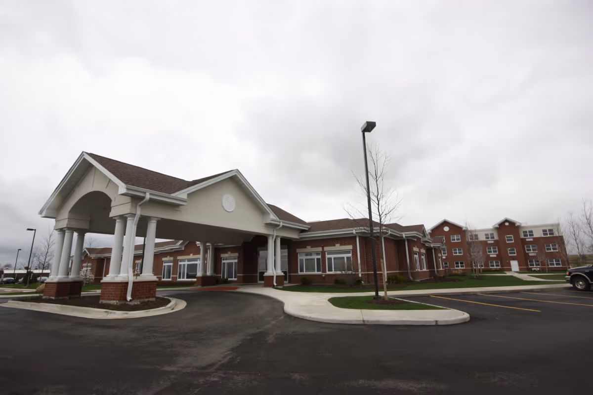 Front entrance and porte-cochère of a red-brick senior living facility with a parking lot under a cloudy sky.