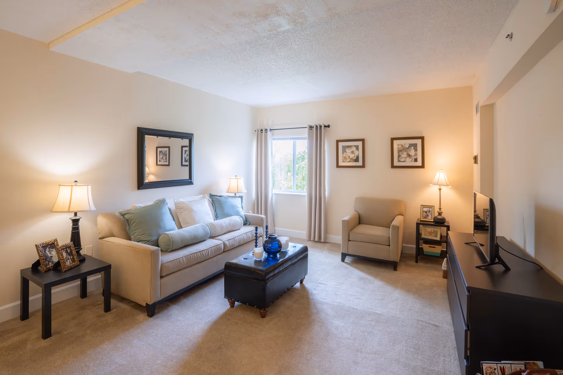 A cozy living room with a beige sofa adorned with blue and white pillows, a matching armchair, a black ottoman coffee table with decorative items, two side tables with lamps, framed pictures on the walls, and a window with light curtains letting in natural light.