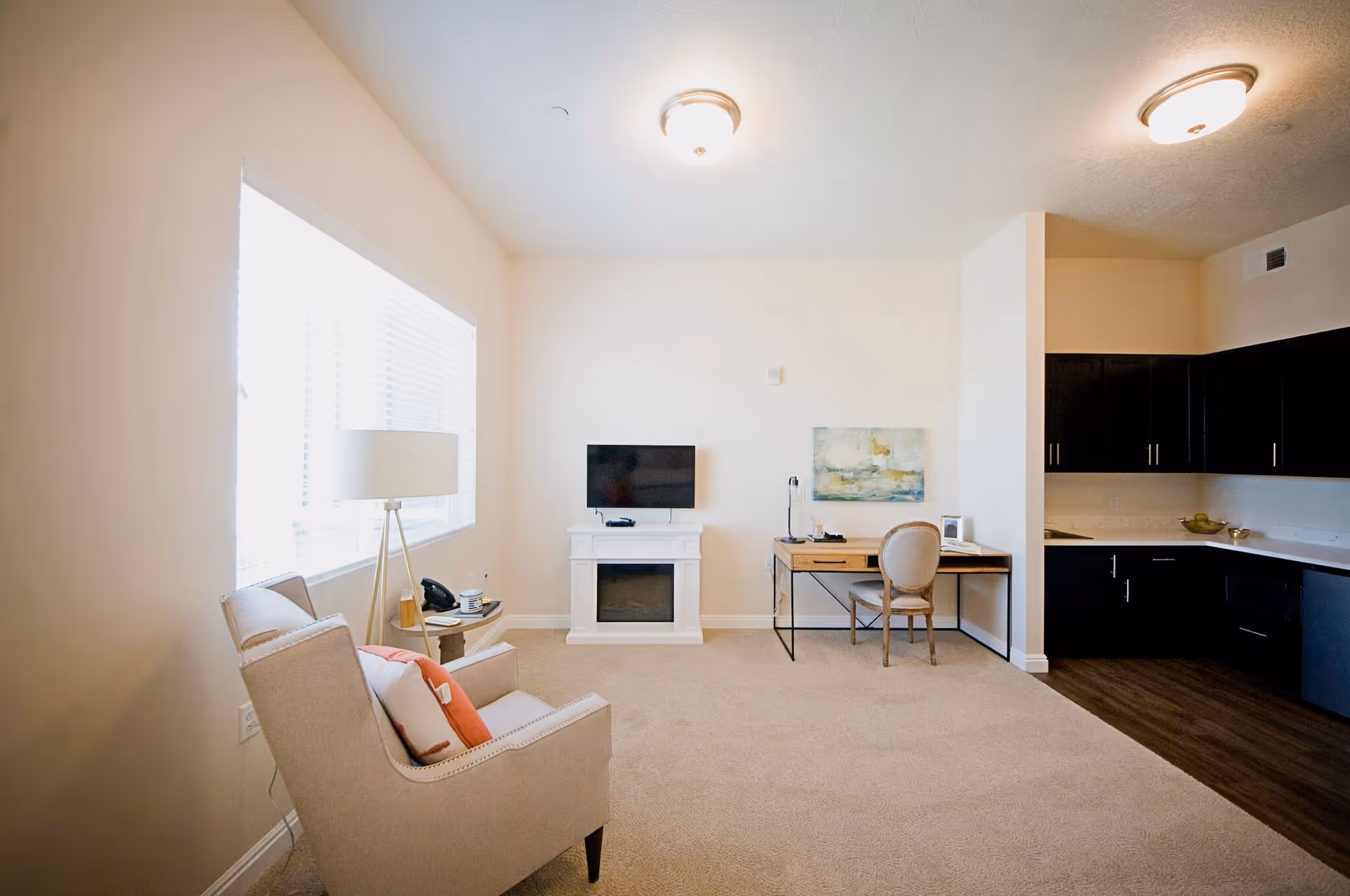 A bright living area with a beige armchair and a floor lamp near a large window on the left. A small white fireplace with a flat-screen TV mounted above it is centered on the far wall. To the right, there is a wooden desk with a chair, a desk lamp, and a piece of artwork hanging above. The room has beige carpet and leads into a kitchen area with dark cabinets and a wooden floor.