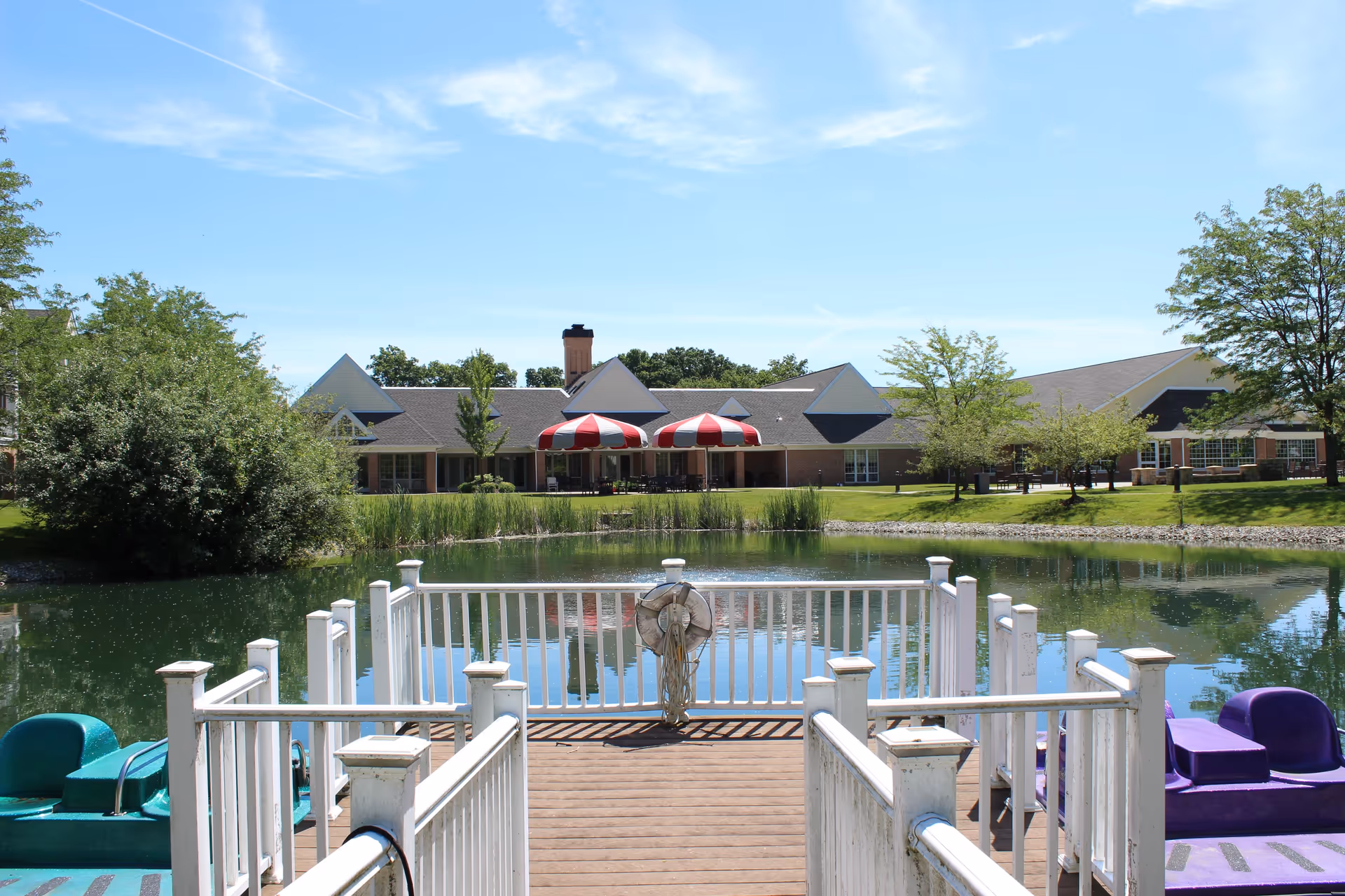 View from a wooden dock with white railings overlooking a small pond with paddle boats on either side. Across the pond is a building with a sloped roof, red and white striped umbrellas, and surrounded by green trees and grass under a blue sky with some clouds.