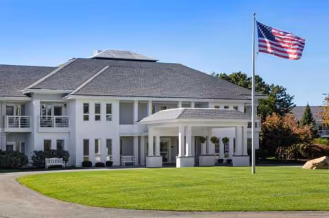 White senior living facility with porte-cochere entrance, green lawn, and American flag.