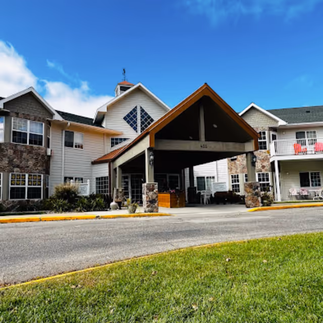 Covered main entrance and porte-cochère of a multi-story senior living building with stone accents, balconies, and a blue sky above.