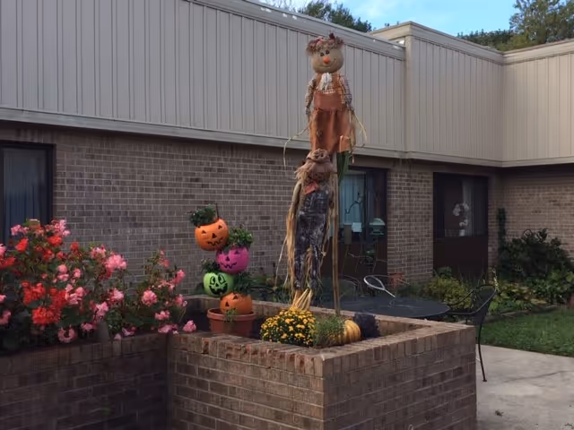 Outdoor courtyard area with brick planters containing pink flowers and Halloween decorations including a tall scarecrow and stacked pumpkin pots with faces. There is a round metal table with chairs and a brick building with windows in the background.