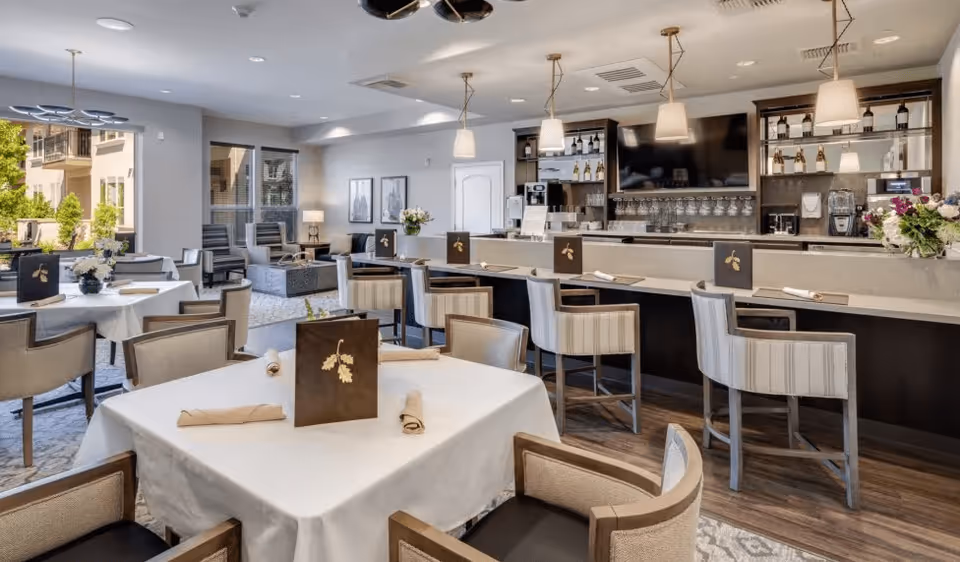 A bright and elegant dining area in a senior living facility featuring tables covered with white tablecloths, each set with rolled napkins and menus. The room includes a bar with high chairs, pendant lighting, and shelves stocked with bottles and glassware. Large windows allow natural light to fill the space, and there is a cozy seating area with armchairs near the windows.