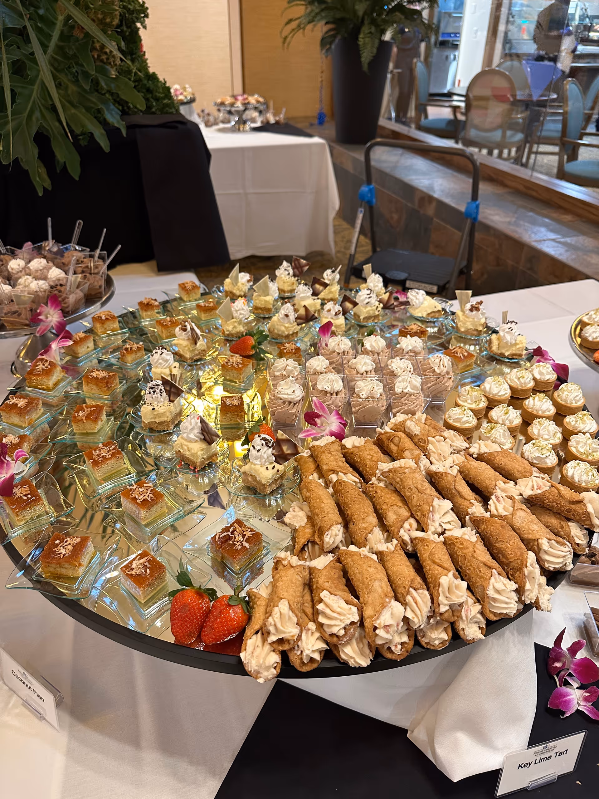 A table in an indoor dining area filled with assorted desserts—cannoli, mini cakes, and mousse cups—arranged on a mirrored serving platter.