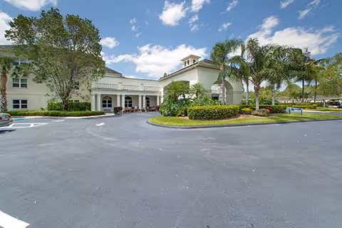 Exterior view of a senior living facility with a circular driveway, palm trees, and well-maintained landscaping under a partly cloudy sky.