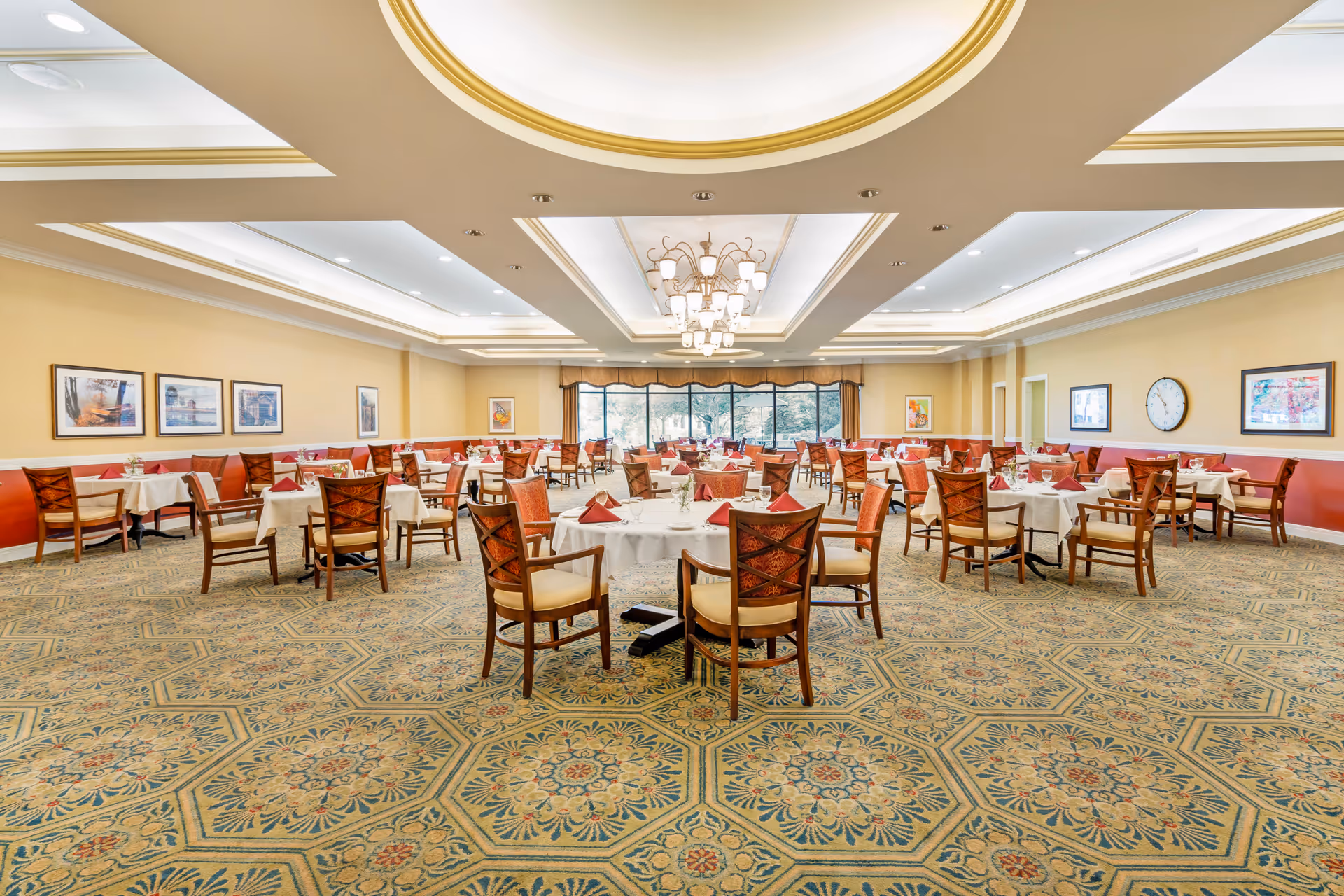 A spacious dining room with multiple round and square tables covered with white tablecloths and set with glasses, plates, and red napkins. The room features a patterned carpet, beige walls with framed artwork, large windows with a view of greenery outside, and a decorative ceiling with recessed lighting and a chandelier.