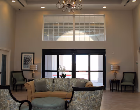 A bright and spacious senior living facility common area with a beige sofa, patterned armchairs, a round ottoman, two side tables with lamps, and a chandelier hanging from the ceiling. Large windows and glass doors allow natural light to fill the room.