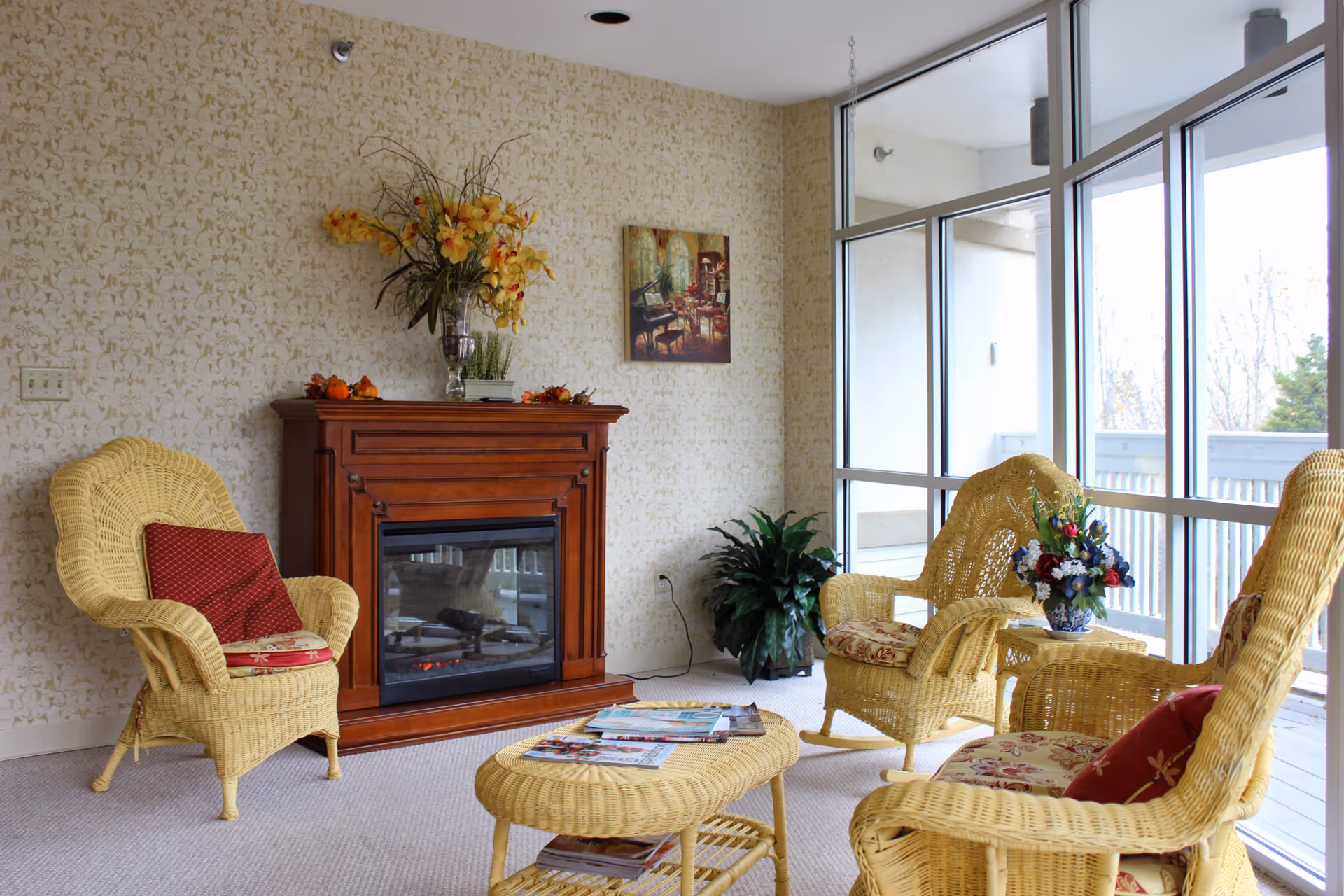 Sunlit seating area with wicker chairs, a wicker coffee table, floral arrangements and an electric fireplace next to large windows.